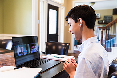 While distance learning during the COVID-19 pandemic, a teenage boy talks with a female teacher via video call. The boy is using a laptop to communicate with the teacher.