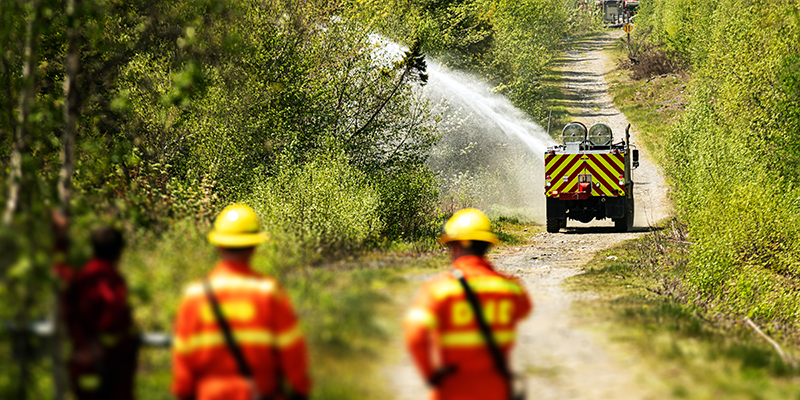 Firefighters watch as an off-road firetruck sprays the edge of a dirt road near a forest fire.